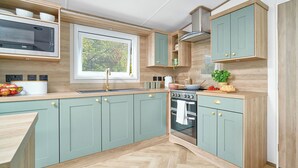 A modern kitchen with light green cabinets, a built-in microwave, a window with blinds, a stove, and a sink. - Diamond 2 Bed Caravan Hot Tub (Clarach Bay, Nr Aberystwyth)