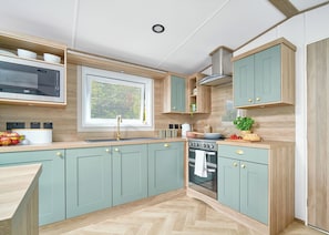 A modern kitchen with light green cabinets, a built-in microwave, a window with blinds, a stove, and a sink. - Diamond 2 Bed Caravan Hot Tub (Clarach Bay, Nr Aberystwyth)