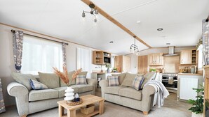 A spacious living room with two sofas, a coffee table, and a kitchen area with wooden cabinets and a stove. - Diamond 2 Bed Caravan Hot Tub (Clarach Bay, Nr Aberystwyth)