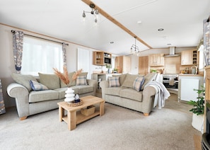 A spacious living room with two sofas, a coffee table, and a kitchen area with wooden cabinets and a stove. - Diamond 2 Bed Caravan Hot Tub (Clarach Bay, Nr Aberystwyth)