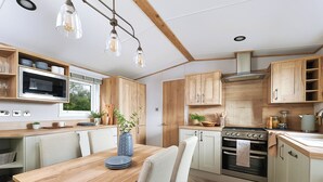 A modern kitchen with wooden cabinets, a built-in oven, and a dining area with a table and chairs. - Diamond 2 Bed Caravan Hot Tub (Clarach Bay, Nr Aberystwyth)