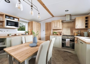 A modern kitchen with wooden cabinets, a built-in oven, and a dining area with a table and chairs. - Diamond 2 Bed Caravan Hot Tub (Clarach Bay, Nr Aberystwyth)
