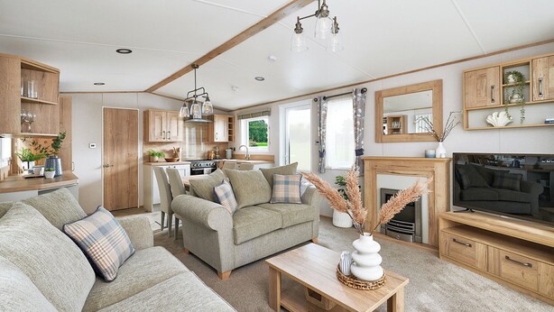 A cozy living room with a sofa, a coffee table, and a TV. There is a kitchen area with wooden cabinets and a dining table. - Diamond 2 Bed Caravan Hot Tub (Clarach Bay, Nr Aberystwyth)