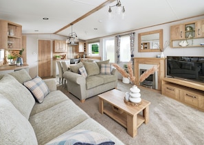 A cozy living room with a sofa, a coffee table, and a TV. There is a kitchen area with wooden cabinets and a dining table. - Diamond 2 Bed Caravan Hot Tub (Clarach Bay, Nr Aberystwyth)