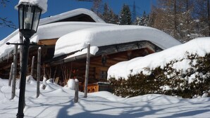 Una baita di legno coperta di neve, con un camino ben visibile, circondata da alberi carichi di neve e un cielo azzurro terso.