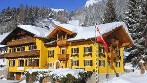 Un edificio giallo con balconi e una bandiera rossa, circondato da alberi e montagne innevate.