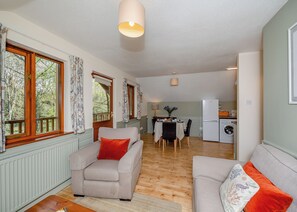 Una sala de estar acogedora con sofá, una mesa de comedor con sillas, una zona de cocina con refrigerador y lavadora, y ventanas amplias con cortinas.