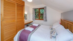 A bedroom with a wooden wardrobe, a bed with a purple and white bedspread, a bedside table with a lamp, and a window with curtains.
