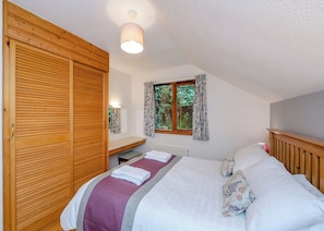 A bedroom with a wooden wardrobe, a bed with a purple and white bedspread, a bedside table with a lamp, and a window with curtains. - Timber lodge set by the lake with one double and one twin. (Webbery, Bideford)