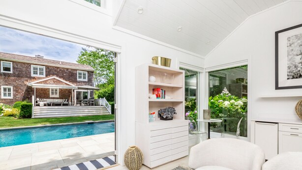Una sala moderna con vista a la piscina, muebles blancos y una puerta de vidrio que da al exterior.