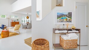 Un amplio espacio habitable con un sofá, una silla y una cesta de mimbre. Se ve una escalera que lleva a un dormitorio con una puerta y un cuadro en la pared.