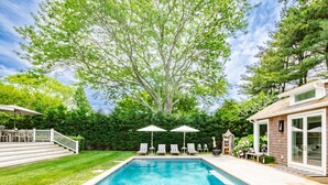 Piscina con sillones de descanso y sombrillas, rodeada de un césped cuidado y árboles.