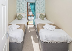 A room with two beds, a small table, a window with curtains, and a view of the outside.
