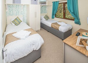 A bedroom with two beds, a wardrobe, a window with curtains, and a small table with a remote control.