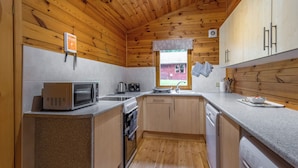A compact kitchen with wooden ceiling, a microwave, a kettle, a sink, and a window with a view of a red building. - Woodland Birch Lodge (Ellesmere)