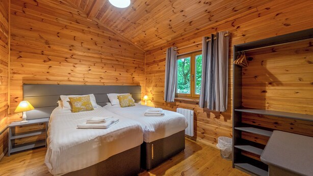 Ein Schlafzimmer in einer Holzhütte mit zwei Betten, einem Fenster mit Vorhängen, einem Nachttisch mit Lampe und einem Sessel.