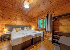 A wooden cabin bedroom with two beds, a window with curtains, a nightstand with a lamp, and a chair. - Woodland Birch Lodge (Ellesmere)