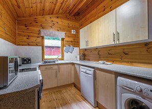 A compact kitchen with wooden cabinets, a dishwasher, a washing machine, and a window with a view of a red building.