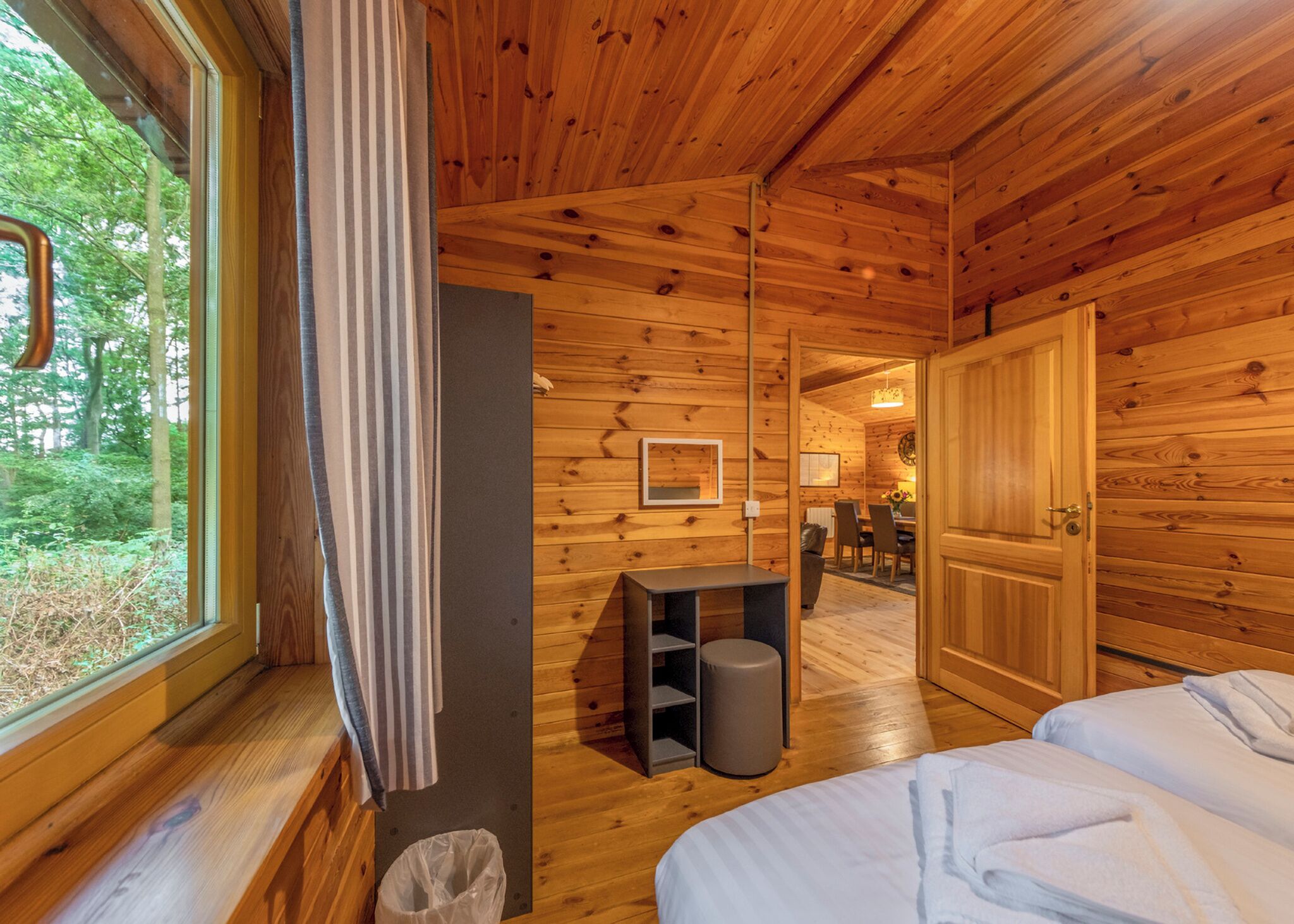 A wooden cabin interior with a bed, a small table, a trash bin, and a door leading to a dining area.