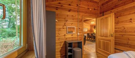 A wooden cabin interior with a bed, a small table, a trash bin, and a door leading to a dining area.
