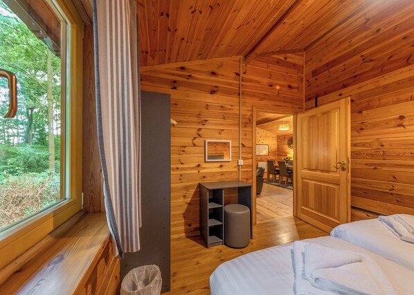 A wooden cabin interior with a bed, a small table, a trash bin, and a door leading to a dining area.