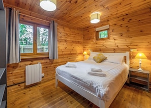 A wooden cabin bedroom with a bed, two bedside lamps, a window with curtains, and a radiator.