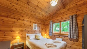A wooden cabin bedroom with a bed, bedside lamps, a window with curtains, and a view of trees. - Woodland Birch Lodge (Ellesmere)