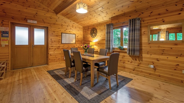 A wooden cabin interior with a dining table, chairs, a clock, and a window with curtains. - Woodland Birch Lodge (Ellesmere)