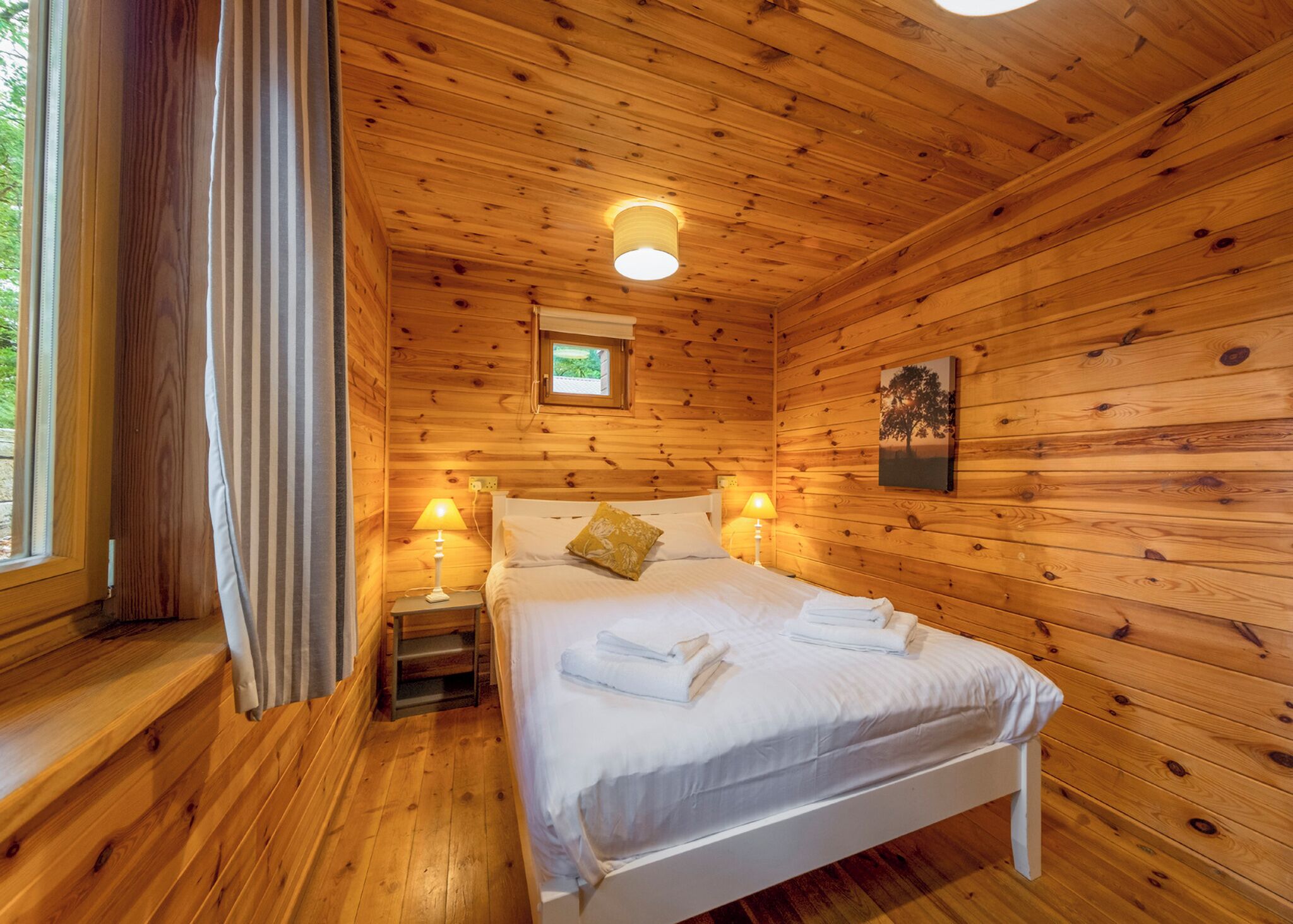 A wooden cabin bedroom with a bed, bedside lamps, a window with curtains, and a small picture on the wall.