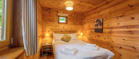 A wooden cabin bedroom with a bed, bedside lamps, a window with curtains, and a small picture on the wall.