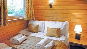 A wooden cabin bedroom with a bed, pillows, a nightstand, a lamp, and a window with curtains. - Timber lodge with one queen size double and one twin. (Kentisbeare, Nr Cullompton)