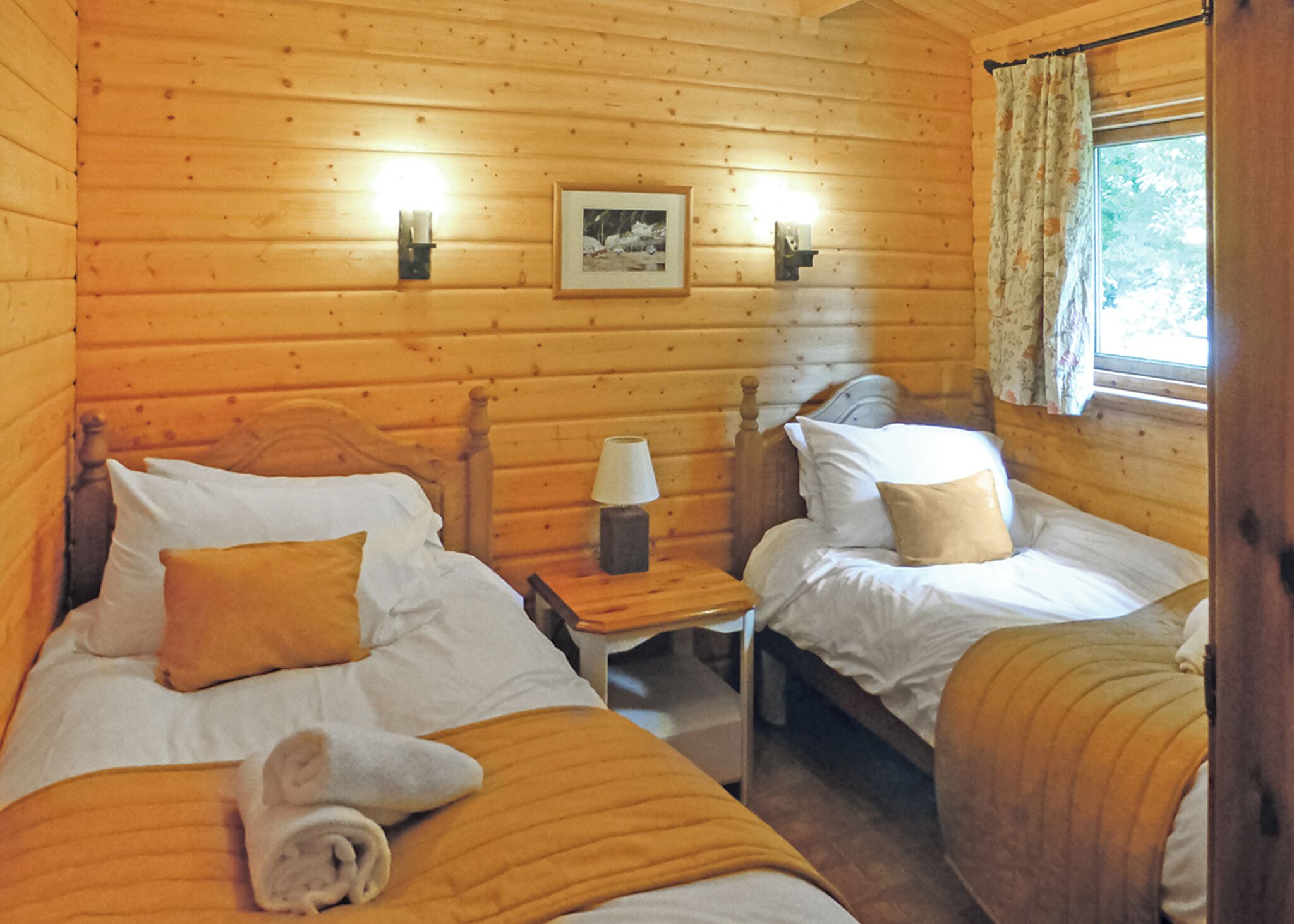 Une chambre de style cabane avec deux lits, une table de chevet avec une lampe, un tableau encadré au mur et une fenêtre avec des rideaux.