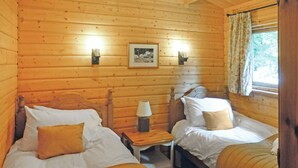 A cabin-style room with two beds, a nightstand with a lamp, a framed picture on the wall, and a window with curtains. - Timber lodge with one queen size double and one twin. (Kentisbeare, Nr Cullompton)