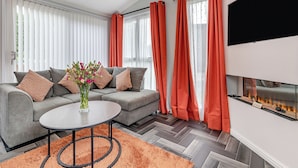 A modern living room with a grey sofa, a round coffee table, a fireplace, and orange curtains. - Buxton Lodge with hot tub (Longnor, Nr. Buxton)