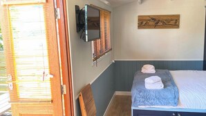 Un dormitorio con cama, una silla de madera, un televisor montado en la pared y una ventana con persianas.