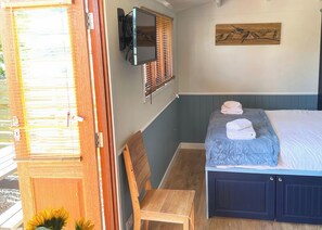 A bedroom with a bed, a wooden chair, a wall-mounted TV, and a window with blinds.