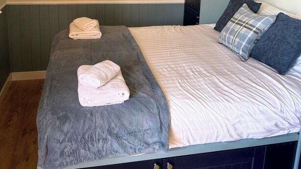 Una cama con dos toallas enrolladas, una manta gris y una colcha blanca con un cojín de cuadros.