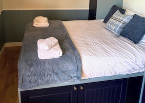 A bed with two rolled towels, a grey blanket, and a white bedspread with a plaid pillow.