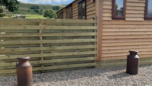 Una cabaña de madera con techo inclinado, cerca de madera y dos contenedores metálicos.