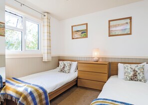 A bedroom with two single beds, a bedside table with a lamp, a window with curtains, and two framed landscape paintings on the wall.