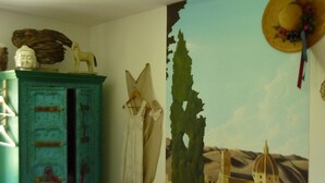 Una habitación con un aparador azul rústico, un vestido blanco en un perchero y un mural de un paisaje con edificios.