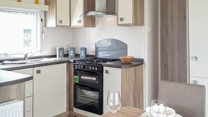 Een kleine keuken met een fornuis, oven en spoelbak, een eettafel met een glas, en een kom op het aanrecht.