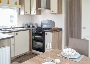 Een kleine keuken met een fornuis, oven en spoelbak, een eettafel met een glas, en een kom op het aanrecht.