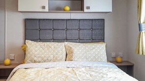 Een bed met een beddekleed met patroon, een grijs gestoffeerd hoofdbord en twee nachtkastjes met gele vazen.