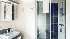 Une salle de bain avec un lavabo blanc, un miroir, une cabine de douche en verre et une serviette accrochée à la porte de la douche.