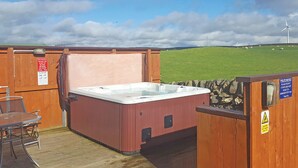 Un bain à remous installé dans une structure en bois à l’extérieur, avec une table et des chaises, un panneau indiquant les heures d’utilisation du bain à remous, et une vue sur un champ verdoyant où se trouve une éolienne.