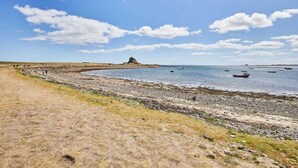 Paisaje costero con una playa arenosa, costa rocosa y una colina distante con una construcción. Se ven barcos en el agua.