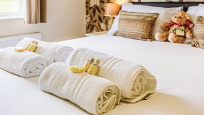 A neatly made bed with rolled white towels and toiletries, a teddy bear, and a lamp on a floral wallpaper wall. - Luxury lodge with king-size bed with en suite shower. (Shrewsbury)