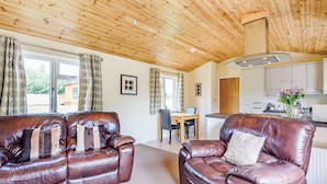 A cozy living area with a wooden ceiling, a brown leather sofa, a dining table with chairs, and a kitchen in the background. - Luxury lodge with king-size bed with en suite shower. (Shrewsbury)