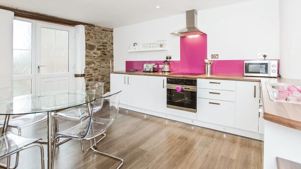 A modern kitchen with a glass dining table, white cabinets, a stone accent wall, and a pink backsplash.